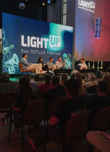 Vier Männer und eine Frau, die nebeneinander auf einem Sofa auf einer Bühne sitzen. Einer hält ein Mikrofon und spricht hinein, die anderen hören zu. Im Hintergrund steht "LightUp Das DOTLUX Festival" auf einer Leinwand.