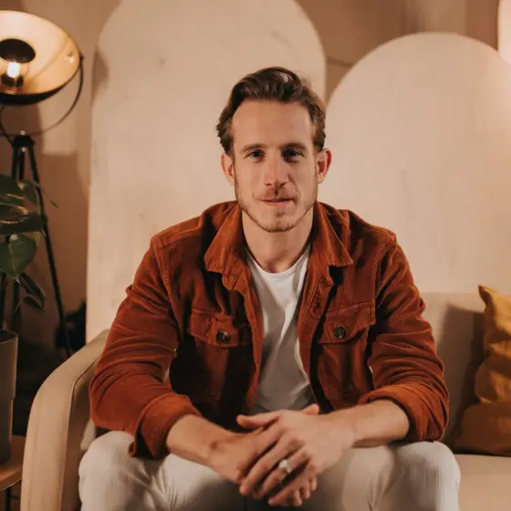 Ein Mann der mit übereinandergelegten Händen auf einem beigen Sofa in einem warm beleuchteten Raum sitzt